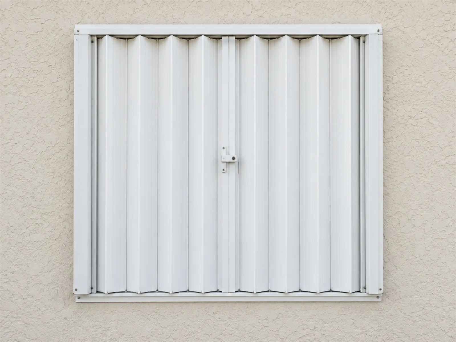 A white accordion shutter closed and latched across a window on a pale stucco wall, its folded slats and centre lock seen straight on in soft daylight